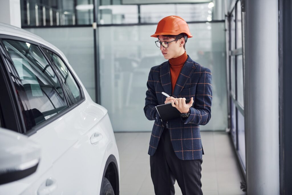 Worker in protective helmet and with notepad looking at modern car